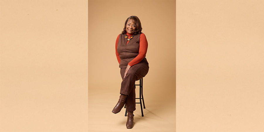Eathell posing for COMP photo shoot. She is a Black woman sitting on a stool with one of her legs crossed over the other. She wears an orange sweater with brown pants and a brown vest.