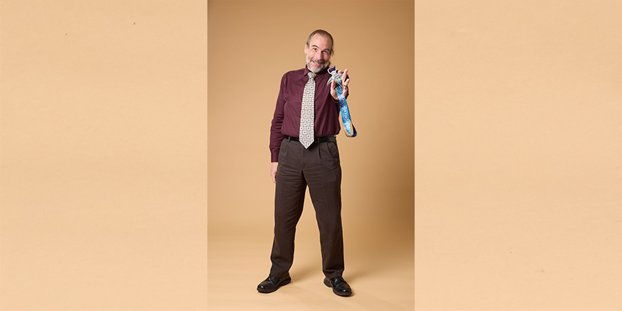 Joe D posing at COMP photo shoot. He is an older white male, dressed in slacks, a nice maroon shirt and tie. He holds a race medal toward the camera.