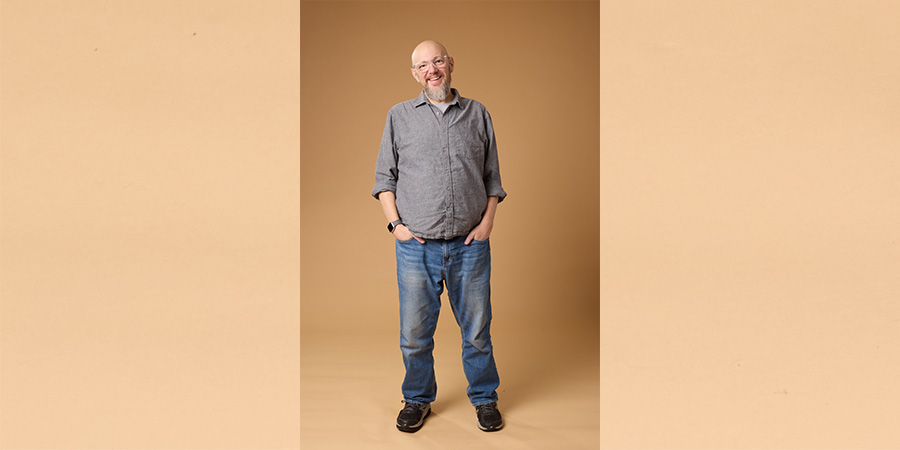 John posing for COMP shoot. He is a white man wearing jeans and a long-sleeve gray button down shirt. 