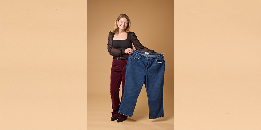Kim poses for COMP photo shoot. She is a short white woman with blond hair, wearing dark red pants and a black top. She holds a pair of pants to her left, stretched wide.