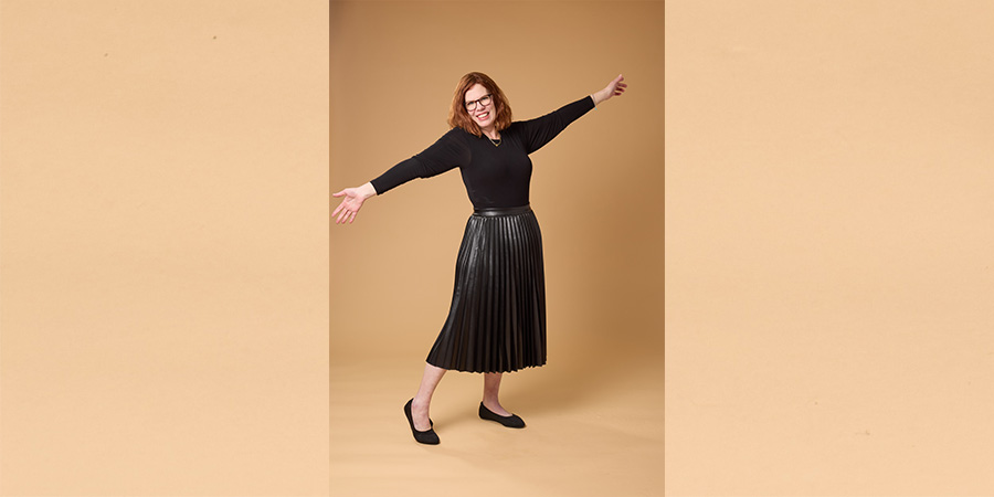 Mel posing on a COMP photo shoot. She is a white woman with red hair, wearing a black shirt and black skirt. 