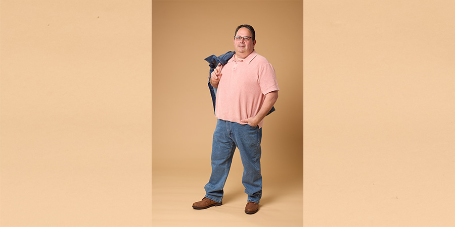 Neal posing for COMP photo shoot. He is a white man wearing jeans and a pink shirt, and holding a jean jacket over his shoulder.