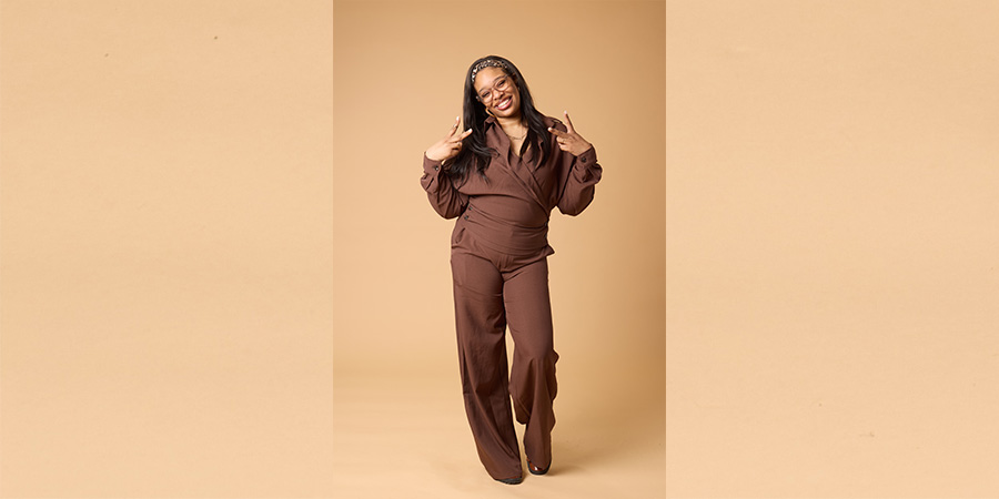 Shana D posing for COMP photo shoot. She is a Black woman posing with the peace sign on both hands, wearing a brown tracksuit.