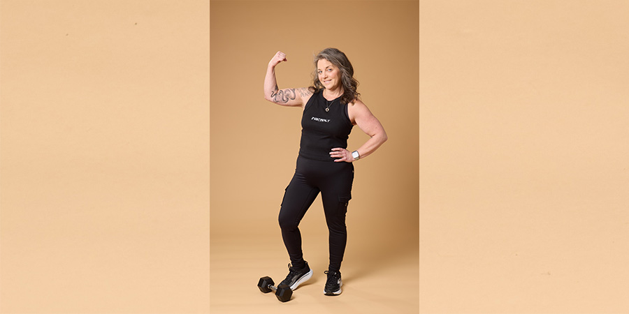 Stacey R poses for COMP photo shoot. She is a white woman in a black workout outfit, flexing for the camera.